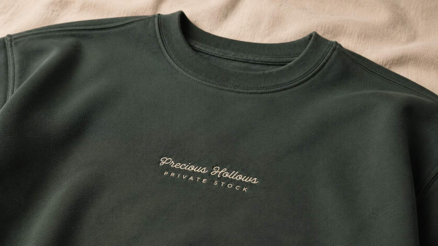 A close-up of a dark forest green crewneck sweatshirt laid flat on a beige fabric background. The chest features a small white embroidered logo that reads 'Precious Hollows PRIVATE STOCK'.