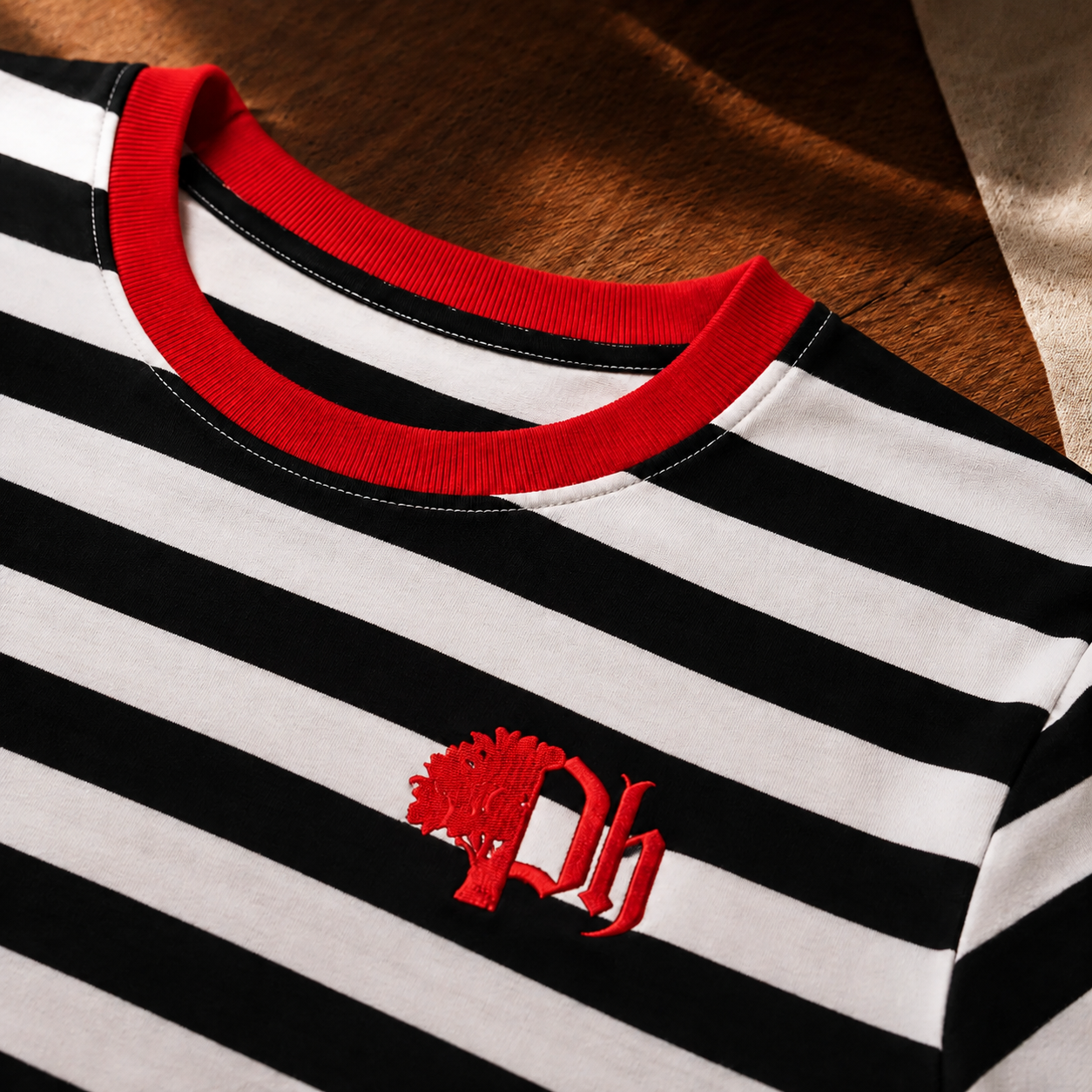 Close-up of a black and white horizontally striped t-shirt with a red ribbed crew neck collar and a red embroidered 'Ph' tree logo on the chest, laid flat on a brown textured surface.