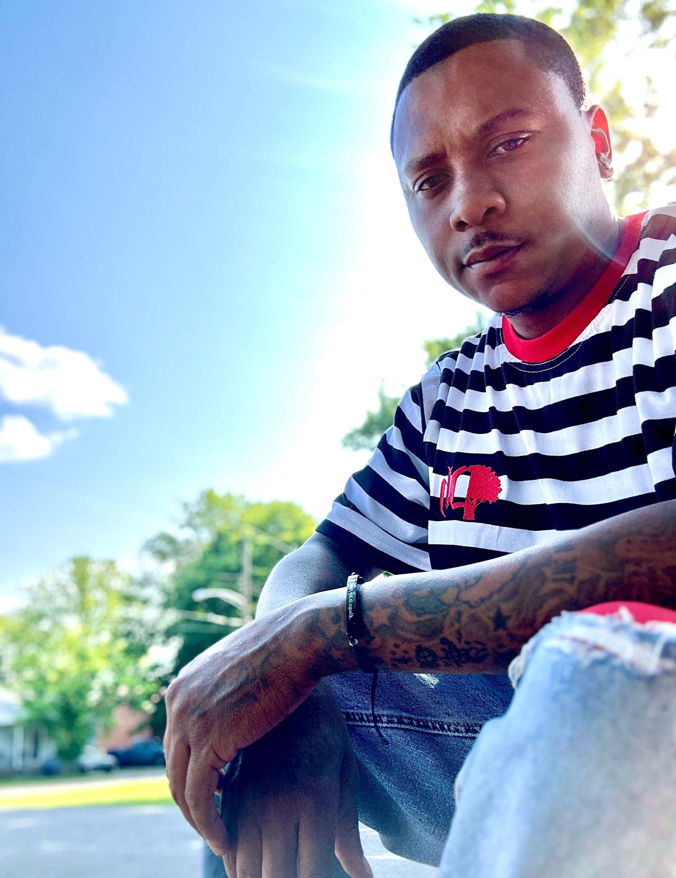 A person wearing a striped cotton t-shirt in a classic fit with a logo on the chest, sitting outdoors.#color_black-stripes
