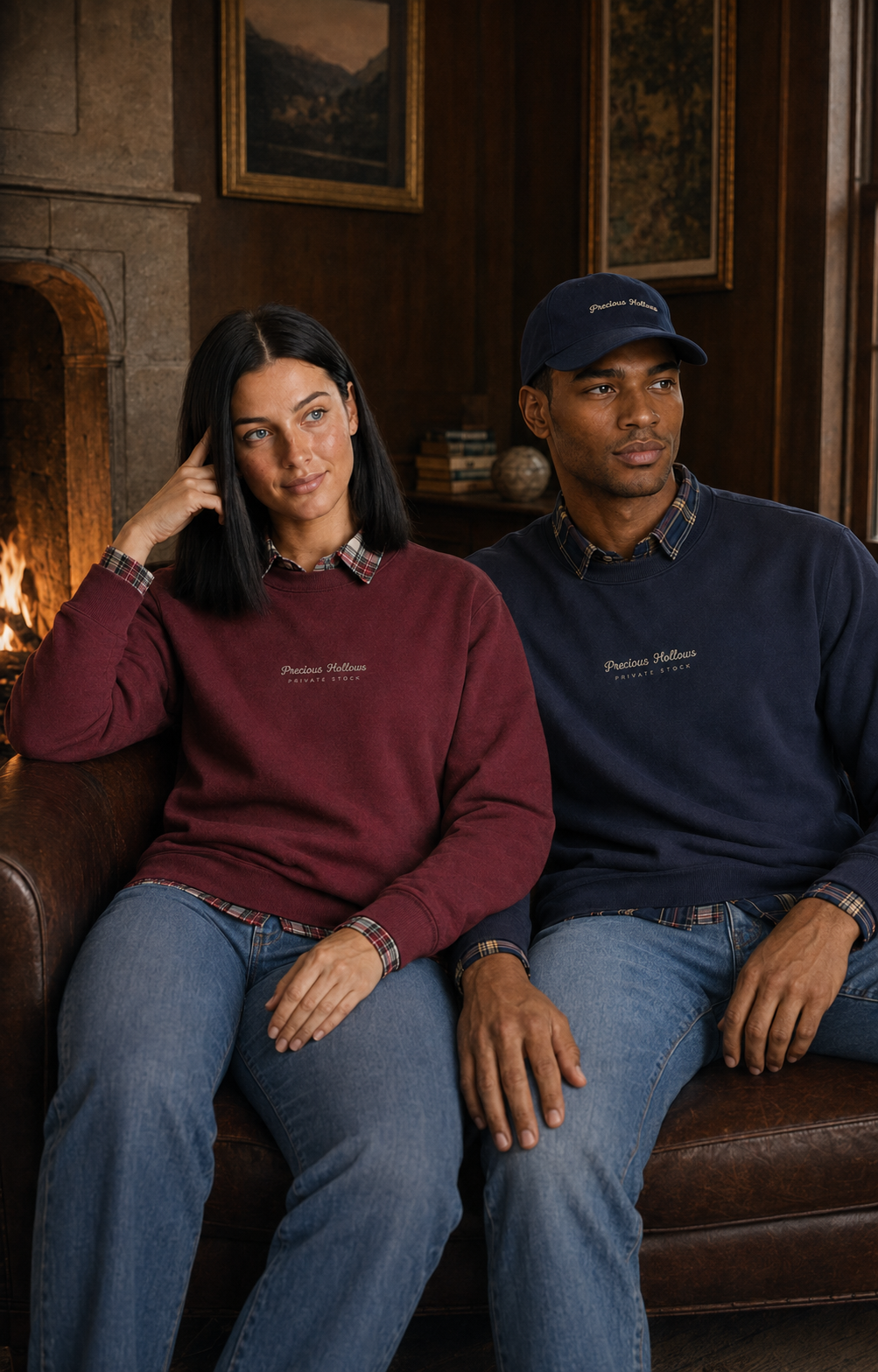 A man and a woman sit side by side on a brown leather couch in a cozy room with a fireplace. Both are wearing matching dark-colored crewneck sweatshirts with 'Pierce Hamilton' embroidered on the chest, paired with blue jeans.