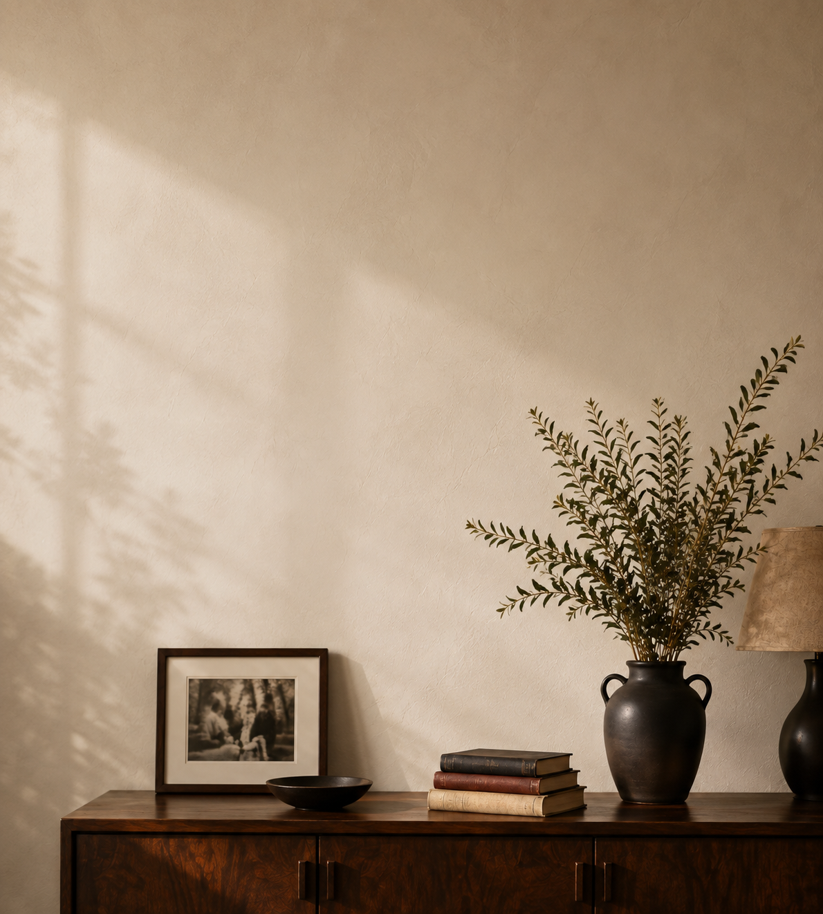 A styled interior scene with a textured beige wall, a dark wood sideboard, a black vase with dried branches, stacked books, a small black bowl, a framed black-and-white photo, and part of a table lamp in the foreground.