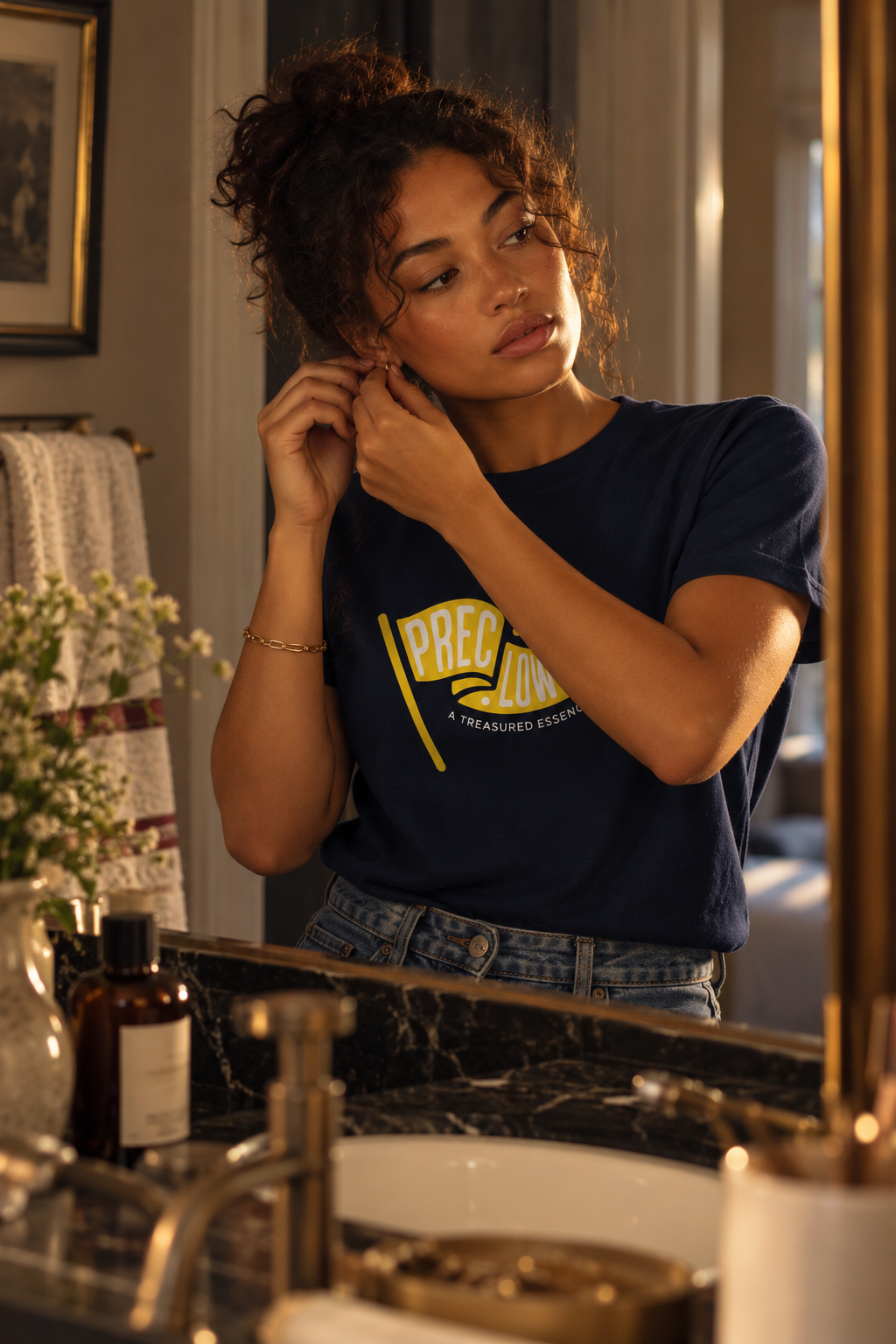 Young woman adjusting an earring at a warmly lit vanity in a navy Precious Hollows tee