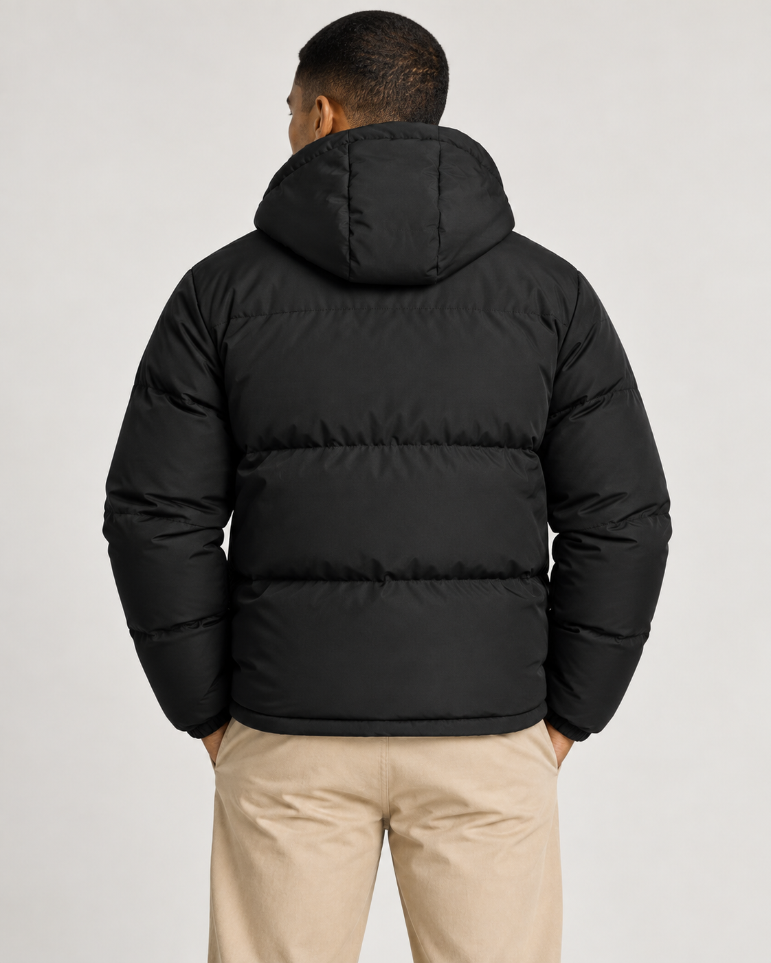 A man viewed from the back wearing a black hooded puffer jacket and beige trousers, standing against a light gray studio background.
