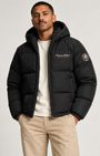 A man wearing a black hooded puffer jacket with 'Precious Hollows PRIVATE STOCK' embroidered on the chest, styled over a cream t-shirt and beige trousers, against a light gray studio background.