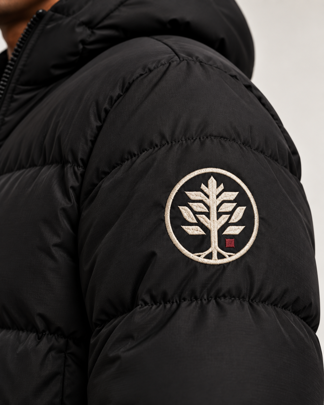 Close-up of a black puffer jacket sleeve featuring a circular embroidered tree logo patch in Off White and Burgundy on the upper arm.