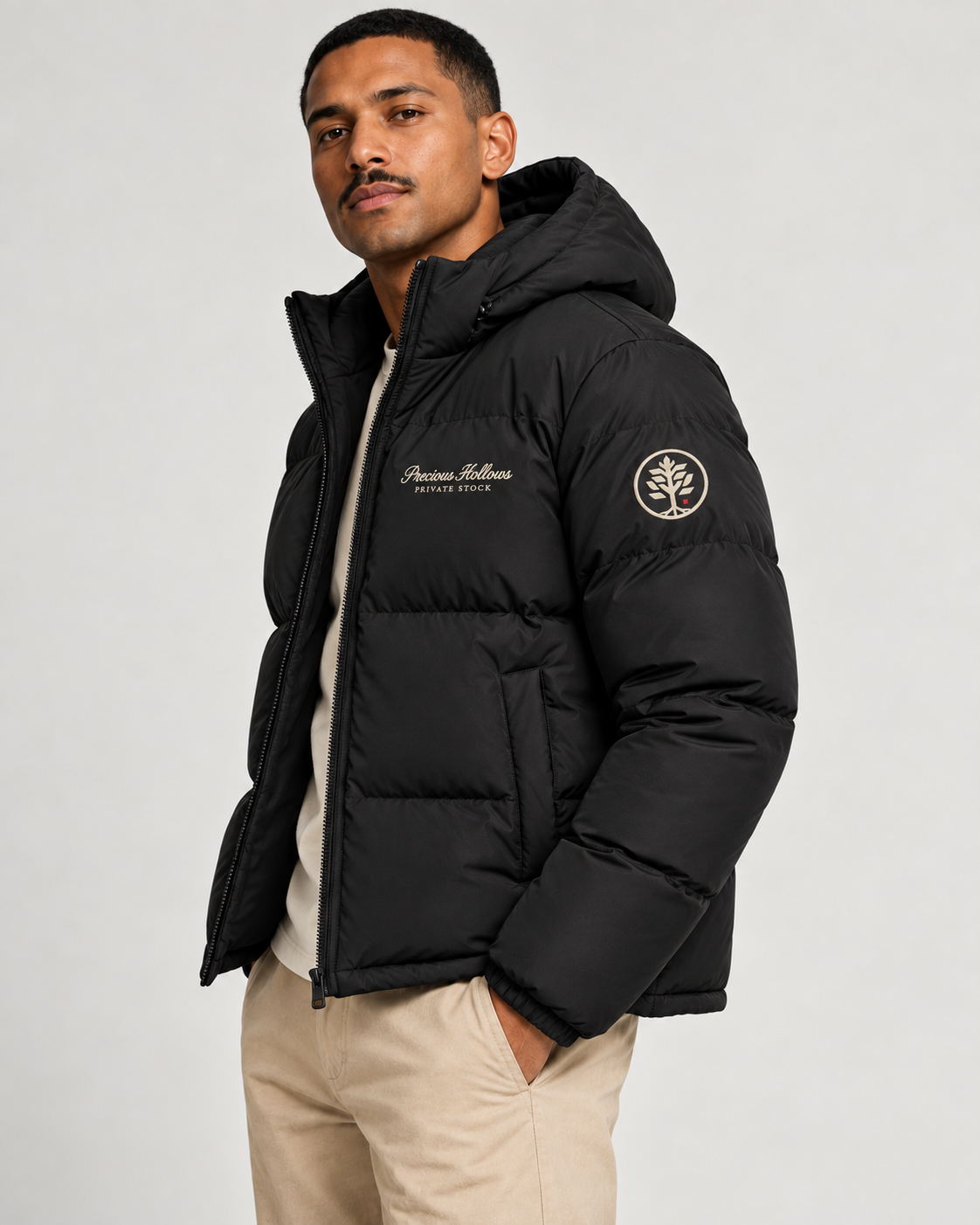 A man wearing a black hooded puffer jacket with Off White embroidered logos on the chest and sleeve, paired with beige pants, against a light gray studio background.