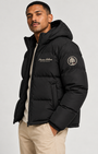 A man wearing a black hooded puffer jacket with Off White embroidered logos on the chest and sleeve, paired with beige pants, against a light gray studio background.