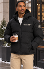 A man wearing a black hooded puffer jacket with 'Precious Hollows PRIVATE STOCK' branding on the chest, standing outdoors in a snowy urban setting and holding a coffee cup.